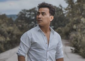 A man in casual white shirt and jeans posing confidently on a rural road with trees around.